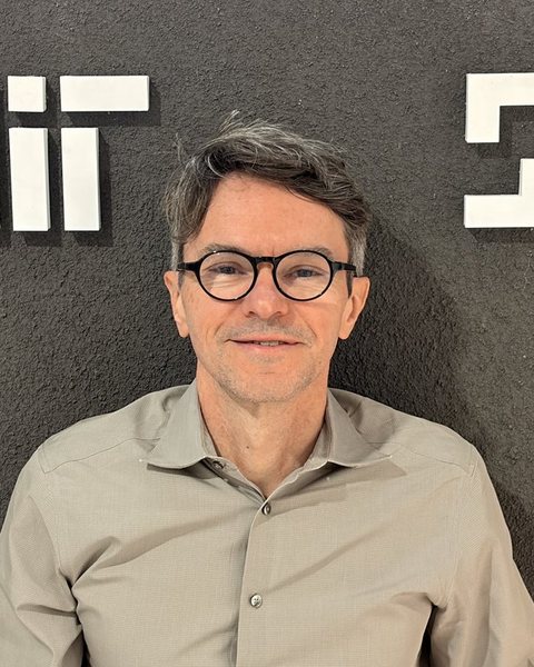 Fabio Duarte in round glasses and an olive shirt, smiling, in front of a dark wall with white MIT lettering.