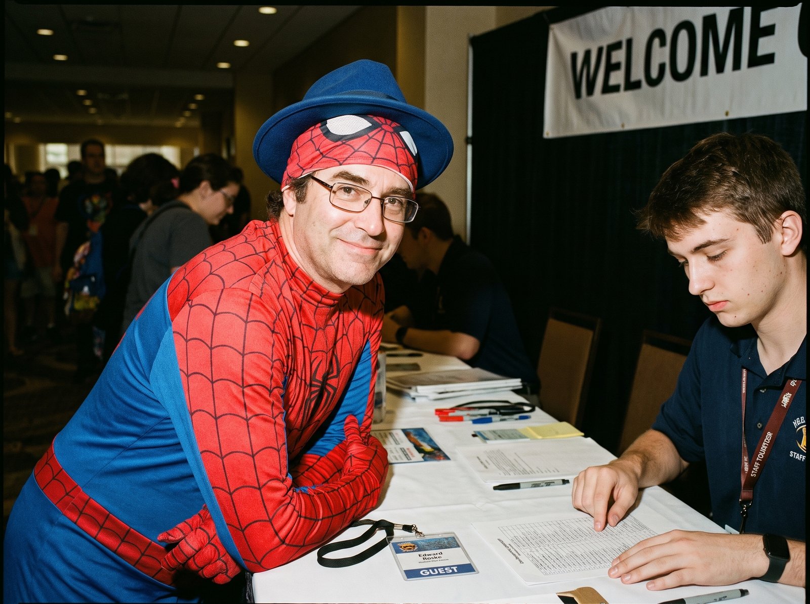 Edward Roske in a red-and-blue web-patterned suit and his blue fedora, leaning politely on a conference registration table while a volunteer in a staff polo scans a printed attendee list without looking up. The badge on the table reads GUEST.