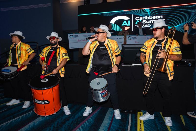 A batucada band in yellow shirts and white hats playing drums and trombone on the main stage