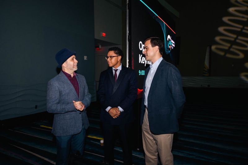 Edward in a blue fedora talking with two men in suits beside a screen that reads Opening Keynote 9:00
