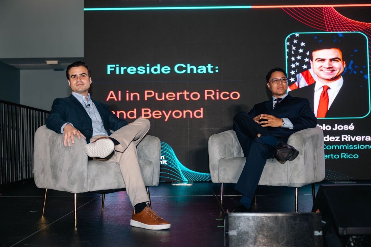 Two men in armchairs on stage under a screen that reads Fireside Chat: AI in Puerto Rico and Beyond