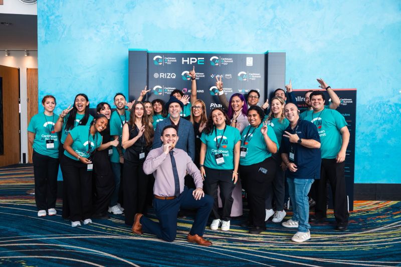 A large group of volunteers in teal Caribbean AI Summit shirts posing in front of the sponsor wall