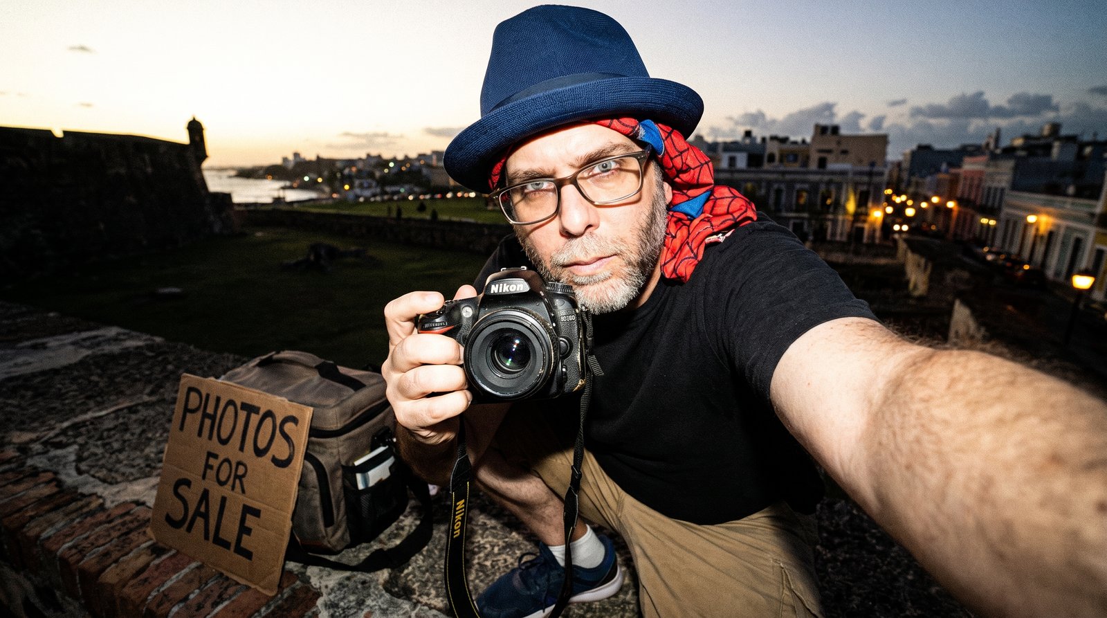 Edward Roske in his signature blue fedora with a red web-patterned superhero mask pushed down around his neck, sitting on the old city wall above Old San Juan at dusk and photographing himself with a battered DSLR held at arm's length, beside a hand-lettered cardboard sign reading PHOTOS FOR SALE.