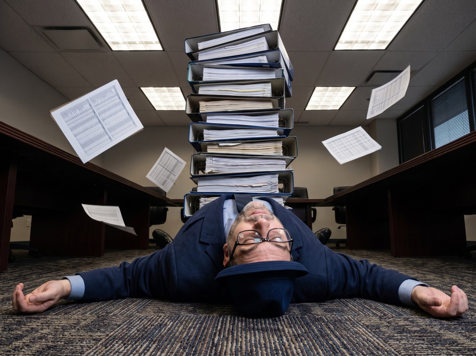 Edward in a navy suit lying flat on his back on a boardroom carpet in slapstick defeat, eyes closed, glasses askew, blue fedora still on his head, under a ceiling-high tower of overflowing accounting binders, spreadsheets fluttering through the air.